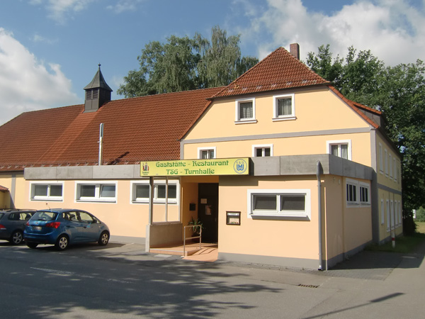 Gelbes Gebäude der TSG-Gaststätte mit Leuchtschild und Vereinslogo an der Fassade