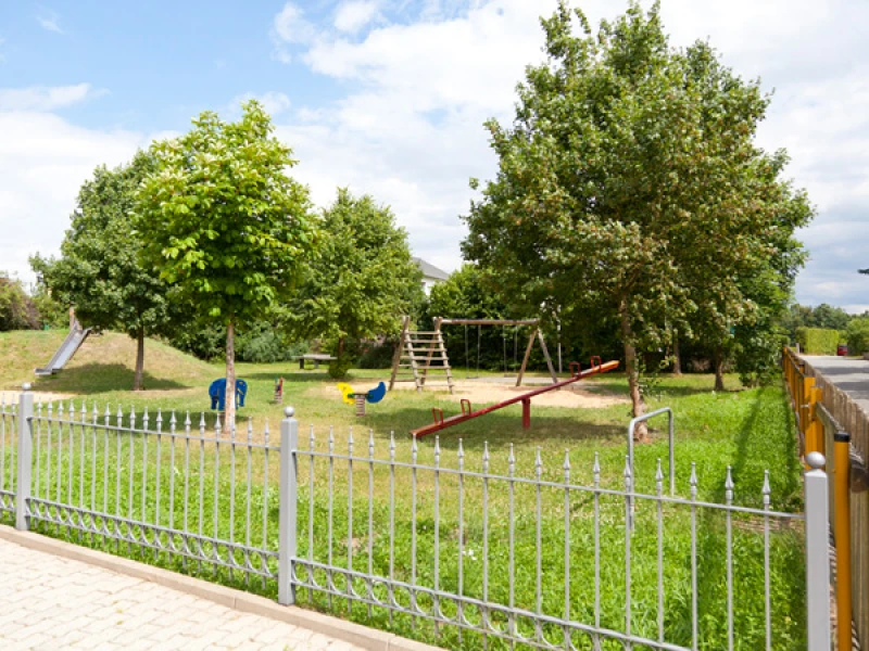 Eingezäunter Spielplatz mit Klettergerüst, Wippe und Rutsche unter Laubbäumen
