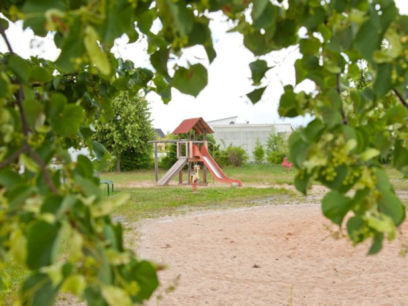 Spielplatz mit Kletterturm, Rutsche und Sandkasten, umrahmt von Baumlaub
